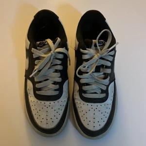 Nike Air Force 1 svart/vit sneakers - Nike Air Force 1 sneakers i svart och vitt skinn med klassisk design. Skorna har perforerad tå, rund tå, platt sula och vita snören. Snygg kontrast mellan svart och vitt, med Nike-logga på hälen och tungan. Perfekta för dig som gillar streetstyle.