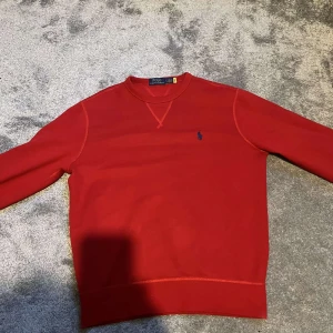 Röd sweatshirt från Polo Ralph Lauren - Riktigt snygg röd sweatshirt från Polo Ralph Lauren med klassisk rund hals och blå broderad logga på bröstet. Tröjan har långa ärmar och ribbade muddar vid ärmslut och nederkant. Perfekt för dig som gillar stilren och enkel design. Knappast använd, Bra skick. 