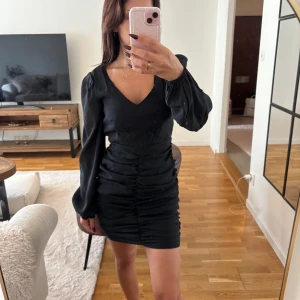 Svart kort omlottklänning NA-KD - Supersnygg svart kort klänning från NA-KD med v-ringning, långa puffärmar och snygg rynkdetalj framtill. Klänningen har en figurnära passform och är gjord i ett glansigt, mjukt material som känns lyxigt. Perfekt för dig som vill sticka ut lite extra!