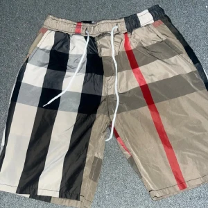 Rutig shorts burrbery  - Snygga shorts med klassiskt rutigt mönster i beige, svart, vit och röd. De har elastisk midja med vit snörning och är tillverkade i lätt polyester. Perfekt för dig som vill sticka ut med en ikonisk streetwear-look.