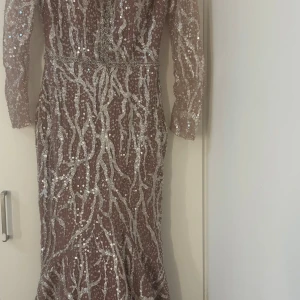Glittrig långklänning med paljetter - Supersnygg långklänning i beige med silverfärgade paljetter och glittrigt mönster. Klänningen har lång ärm, figurnära passform och v-ringning i ryggen. Perfekt för dig som vill sticka ut med en glamorös look.