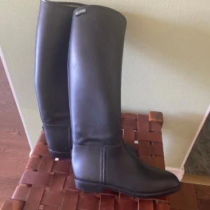 Svarta höga stövlar från Aigle - Snygga svarta stövlar från Aigle med klassisk, stilren design. De är höga med rund tå och platt sula, tillverkade i ett slätt syntetmaterial. Perfekta för dig som gillar minimalistisk look och vill ha ett par boots som funkar året runt.