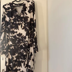 Svartvit blommig omlottklänning - Snygg svartvit klänning med blommigt mönster och omlottmodell. Klänningen har långa ärmar och ett knytband i midjan som ger en fin siluett. Perfekt för dig som gillar stilrena kontraster och en feminin look.