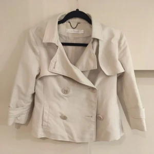 Beige kort trenchcoat - Snygg kort trenchcoat i beige med dubbelknäppning och klassisk krage. Jackan har stora knappar framtill, fickor på sidorna och uppvikta ärmslut. Perfekt för vår och höst💕