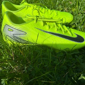 Neongula Nike fotbollsskor - Säljer ett par riktigt snygga neongula Nike fotbollsskor med svart swoosh och silvriga detaljer. Skorna har snörning och är tillverkade i lätt syntetmaterial för bästa känsla på planen. Utrustade med fasta dobbar för gräs. Perfekta för dig som vill sticka ut på matchen!