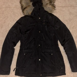 Svart parkasjacka med fuskpäls - Svart parkasjacka från Only med huva och fuskpäls. Jackan har flera fickor med både dragkedja och knappar, samt snörning i midjan för snygg passform. Perfekt för kalla dagar med sin vadderade insida och långa ärmar.