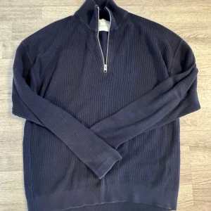 Mörkblå ribbad half zip stickad tröja - Mörkblå stickad tröja med ribbad struktur och half zip-dragkedja i halsen. Långärmad modell med hög krage, perfekt för kyligare dagar. Enkel och stilren design som passar till det mesta. Storlek L