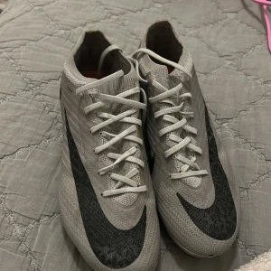 Nike Mercurial Zoom fotbollsskor - Säljer ett par Nike Mercurial Zoom fotbollsskor i ljusgrått med stora svarta swoosh-loggor. Ovandelen är i syntetmaterial med snörning och sulan har rosa detaljer och tydliga dobbar för gräsplan. Perfekta för dig som vill ha snabba och lätta skor på planen storlek 38,5.