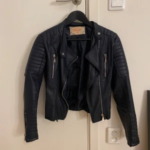 Svart skinnjacka från Vogelle - Snygg svart skinnjacka från Vogelle med coola detaljer som dragkedjor på bröstet och ärmarna samt quiltade partier på axlar och armar. Jackan har en klassisk bikerstil och är perfekt för dig som vill ha en edgy look.