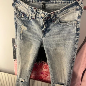 True Religion ljusa bootcut jeans - Snygga ljusblå jeans från True Religion i modellen Becca MidRise Bootcut. Jeansen har slitna detaljer, röda kontrastsömmar och klassiska fickor med lock och knappar bak. Jättefint skick, använts högst 3 gånger. Original pris 1300kr 