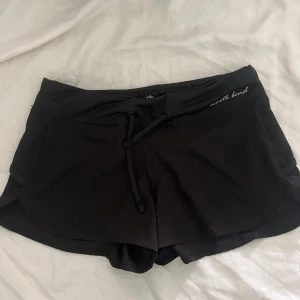 Svarta träningsshorts North Bend - Säljer ett par svarta träningsshorts från North Bend i storlek 34. De har en snygg, sportig design med snörning i midjan och diskret logga på sidan. Materialet är mjukt och syntetiskt, perfekt för aktiva dagar. Passformen är normal och midjan är midwaist.
