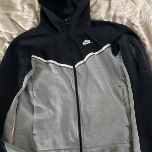 Nike grå och svart tröja S - Nike vindjacka i snygg grå och svart färg med vit logga på bröstet. Jackan har huva, hel dragkedja framtill och en extra ficka med dragkedja på ärmen. Sportig design med kontrastpaneler och vita detaljer längs bröstet. Perfekt för chill eller träning.