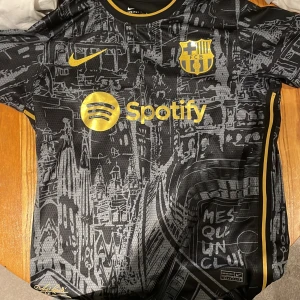 FC Barcelona Lamine Yamal Nike tröja S - Snygg FC Barcelona fotbollströja med Lamine Yamal och nummer 19 på ryggen. Svart bas med guldiga detaljer, klubbmärke och sponsortryck. Unikt grafiskt mönster inspirerat av Barcelonas stadssilhuett. Tillverkad i Nike Dri-FIT material för bästa komfort.