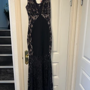 Svart spetsklänning från Lipsy London - Supersnygg långklänning från Lipsy London i svart med transparent spets och blommigt mönster. Klänningen har figurnära passform, är ärmlös och har v-ringad hals. Materialet är polyester med spetsdetaljer som ger en lyxig känsla. Perfekt för att sticka ut med en elegant look.