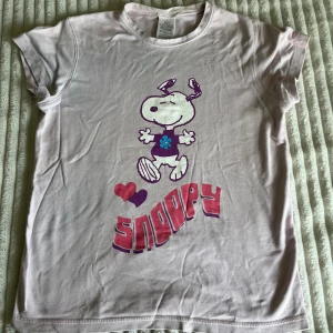 Snoopy baby tee från H&M, lila/rosa - Lila/rosa t-shirt med korta ärmar, prydd med en stor Snoopy-tryck i lila och blått samt texten 'Snoopy' i rosa och lila. Materialet är mjuk bomull och passformen är normal. Perfekt för dig som gillar coola och lekfulla prints.