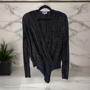 Svart omlott body från Make Way - Jättesnygg svart omlott body från Make Way i storlek S. Bodyn har långärmade ärmar, v-ringning och är sydd i ett glansigt, plisserat tyg med tunna vertikala ränder. 