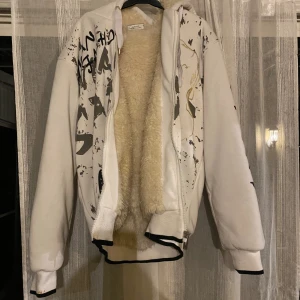 Vit hoodie med tryck och fuskpäls - Vit hoodie med svart och guld grafiskt tryck, dragkedja och huva. Insidan är fodrad med mjuk fuskpäls för extra värme. Den är dock smutsig vid händerna men har tyvärr inget vanish hemma men har tvättats 3 gånger innan men enbart blivit ljusare