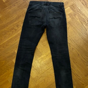 Svarta skinny jeans från Nudie Jeans - Säljer ett par svarta grim tim  från Nudie Jeans i storlek W28 L32. Jeansen har klassisk femficksdesign, svarta sömmar och Nudie-loggan på bakfickan. Snygg tvättad look och smal passform som sitter skönt vid benen. priset kan såklart diskuteras.