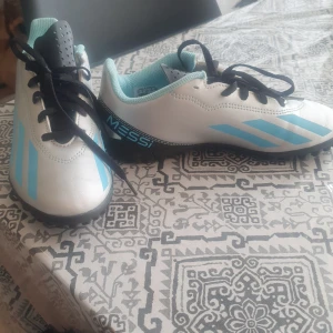 Adidas Messi fotbollsskor strl 36,5 - Säljer ett par nya i silver och ljusblått med svarta detaljer. Skorna har snörning, platt sula med gummidobbar och klassiska Adidas-ränder på sidan. Messi-logga på hälen och insidan. Perfekta för dig som älskar fotboll och vill sticka ut på planen. Aldrig använda.