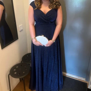 Marinblå glittrig långklänning - Säljer en marinblå balklänninh med glittrigt tyg och v-ringad omlott överdel. Klänningen har korta ärmar och ett elegant fall som ger en lyxig känsla. Använd 2 gånger!💙
