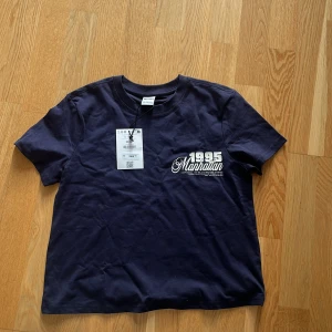 Mörkblå Manhattan t-shirt från Bershka - Snygg mörkblå t-shirt från Bershka med tryck '1995 Manhattan' i vitt på bröstet. Klassisk rund hals och korta ärmar. Perfekt för dig som gillar streetstyle och enkel design.