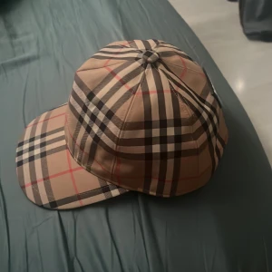 Burberry rutig keps i beige och svart - Snygg keps från Burberry med klassiskt rutigt mönster i beige, svart, vitt och röda linjer. Märket 'BURBERRY LONDON ENGLAND' syns tydligt baktill. Kepsen har böjd skärm och justerbar rem. Perfekt för dig som vill ha en lyxig streetstyle-accessoar.