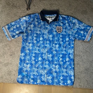 England retro fotbollströja  - Retroinspirerad England fotbollströja från Score Draw med blått geometriskt mönster, klassisk krage i mörkblått och vita detaljer. Broderat England-emblem på bröstet. Tillverkad i lätt och ventilerande material, perfekt för match eller samling.