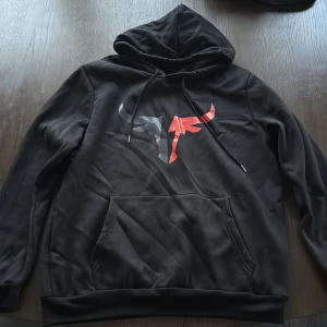 Svart hoodie med grafiskt tryck - Svart hoodie med stor huva och dragsnören. På bröstet finns ett grafiskt tryck i rött och grått som föreställer en stiliserad tjur. Tröjan har en klassisk känguruficka framtill och är tillverkad i mjukt material, perfekt för chill dagar.