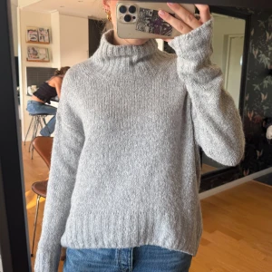 Grå polotröja i stickad ull - Mysig grå polotröja med ribbad krage och muddar. Stickad i mjuk ull, perfekt för kyliga dagar. Tröjan har en loose passform och långa ärmar som ger en chill vibe. Enkel att matcha med jeans eller andra favoritplagg.