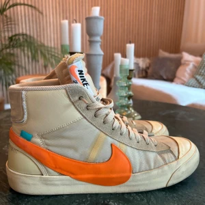 Nike Blazer Mid x Off-White Beige/Orange - Nike Blazer Mid i samarbete med Off-White. Beige sneakers med tydliga orange swoosh, textdetaljer på sidan och klassisk hög siluett. Ovandel i skinn och textil, snörning framtill och Off-White signaturdetaljer. Perfekt för dig som gillar unika och hypade sneakers.