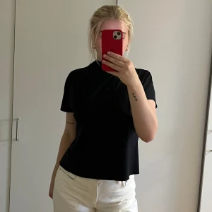 Svart polotopp i mjukt material - Säljer en svart polotopp med korta ärmar och hög krage. Toppen har en rak passform och är tillverkad i ett mjukt, stretchigt material som känns skönt mot huden. Perfekt att styla med jeans eller kjol för en clean look. Använd ett fåtal gånger