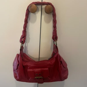 Röd axelväska med flätat handtag - Cool röd axelväska i skinn med flätat handtag och snygga metalldetaljer. Väskan har flera fickor med lock och dragkedja upptill. Perfekt storlek för att få plats med allt du behöver och ger en riktigt trendig vibe. Den rengörs innan skickad!