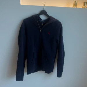 Marinblå hoodie från Polo Ralph Lauren - Snygg marinblå hoodie från Polo Ralph Lauren i storlek XL i barnstorlek motsvarar ( 163-174 cm). Tröjan har dragkedja framtill, huva och två fickor. Klassisk röd broderad logga på bröstet. Perfekt för en chill och stilren look. Säljer pga förliten