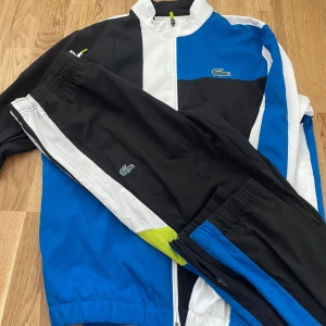 Blå, svart och vit Lacoste sportdress - Sportig dress från Lacoste i blått, svart och vitt med dragkedja framtill och stor krokodil-logga på ryggen. Dressen har meshfoder, ståkrage och coola färgblock. Perfekt för dig som gillar streetwear och sportig stil.