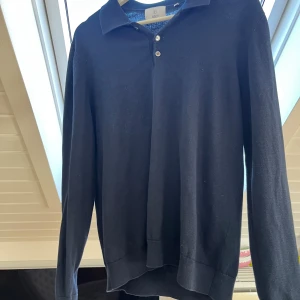 Mörkblå  pikétröja från Bläck, M - Klassisk mörkblå  pikétröja från Bläck i storlek M. Tröjan har lång ärm, krage och tre knappar framtill. Tillverkad i mjukt bomullsmaterial som känns skönt mot huden. Perfekt för en clean och stilren look. Storleken är liten så passar nog en s