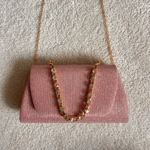 Glittrig rosa kuvertväska med kedja - Säljer en glittrig rosa kuvertväska med guldfärgad kedja och dekorativt handtag med stora stenar. Väskan har en elegant och stilren form, perfekt för att lyfta din outfit. Materialet är syntet med glittrig finish och väskan stängs med lock. Köpt i Italien så minns inte vad affären hette 