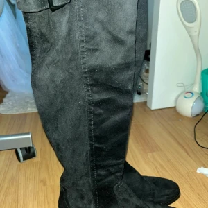 Svarta höga mockastövlar - Snygga svarta stövlar i mocka med högt skaft som går över knäet. De har en enkel design med rund tå och platt sula, perfekta för att lyfta vilken outfit som helst. Stövlarna har en diskret söm längs sidan och en liten detalj upptill.