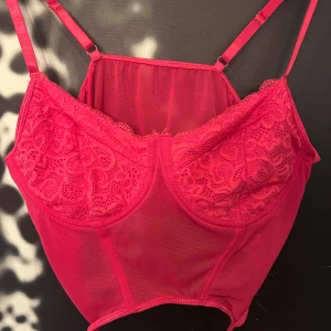 Rosa halv spetskorsett från SHEIN - Säljer en rosa halv korsett-topp från SHEIN med tunna axelband, spetsdetaljer över bysten och mesh-tyg över resten av toppen. Toppen har snörning nertill och är croppad för en trendig look. Perfekt för dig som vill sticka ut med en feminin vibe. Till en kavaj eller under en annan blus då denna blir en snygg detalj 