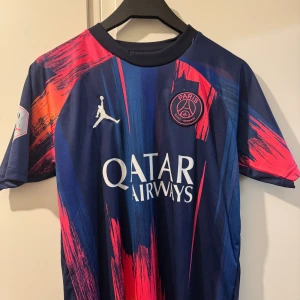 PSG Mbappé Jordan fotbollströja blå/röd - Snygg PSG fotbollströja med Mbappé 7 på ryggen, designad i samarbete med Jordan. Tröjan har ett unikt blått och rött mönster med vita detaljer och klubbmärke på bröstet. Tillverkad i lätt och ventilerande material, perfekt för match eller träning.