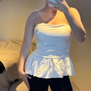 Vit volang tubtopp med peplum - Säljer denna så söta och snygga  vit axelbandslös tubtopp med volangkant nertill och peplumformad siluett. Toppen har en lätt rynkad effekt över bysten och midjan för en snygg passform. Perfekt för dig som vill ha en stilren och trendig look! 😍✨Prislapp kvar! ❤️