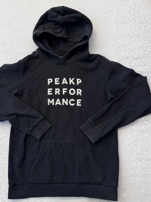 Svart hoodie från Peak Performance - Svart hoodie från Peak Performance med vit text på bröstet. Klassisk känguruficka framtill, ribbade muddar vid ärmslut och nederkant samt stor huva. Tillverkad i mjukt bomullsmaterial som känns skönt mot huden. Perfekt för en avslappnad och sportig stil.