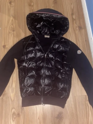 Svart dunjacka med huva från Moncler - Snygg svart dunjacka från Moncler med glansig quiltad front och huva. Jackan har dragkedja framtill, Moncler-logga på ärmen och mjuka ärmar i kontrasterande material. Perfekt för kalla dagar och riktigt stilren look. Priset kan diskuteras vid snabb affär