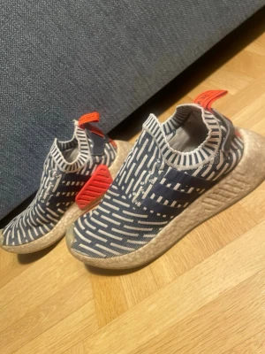 Adidas NMD - Säljer ett par Adidas NMD City Sock sneakers i blått och vitt med stickad ovandel och grafiskt mönster. Skorna har elastisk strumpliknande passform, vita Boost-sulor och röda detaljer på häl och yttersula. Perfekta för dig som gillar streetwear och unik design.