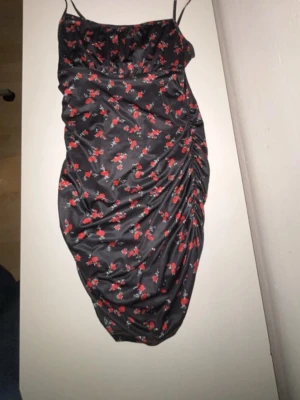 Svart blommig kort klänning - Snygg svart kort klänning med röda blommor och smala axelband. Klänningen har rynkade detaljer på sidan och sitter tight. Perfekt för dig som gillar en feminin och trendig stil.