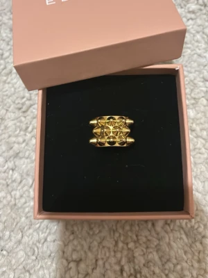 Guldfärgad ring från Edblad - Säljer en statementring från Edblad i guldfärgad metall. Ringen har en bred design med upphöjda geometriska former som ger en cool och modern look. Perfekt för dig som vill sticka ut med dina accessoarer.