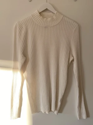 Vit ribbad polotröja från qarma  - Säljer en stilren vit ribbstickad polotröja med lång ärm och hög krage. Tröjan har en tight passform och är gjord i ett mjukt, stretchigt material som sitter snyggt på kroppen.