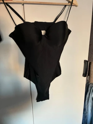 Svart body med tunna axelband - Snygg svart  body  med tunna axelband och vadderade kupor. Klassisk modell med liten cut-out framtill som ger en modern touch. Tillverkad i stretchigt material som sitter skönt mot kroppen.  Storlek 80c, endast använd en gång 