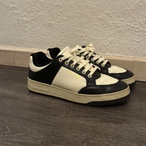Saint Laurent SL61 sneakers - Svartvita sneakers från Saint Laurent med klassisk låg siluett. Skorna har svart och vit ovandel i skinn och textil, perforerade detaljer och tydlig logga på sidan. Platt sula och snörning framtill ger en clean och tidlös vibe. De är i perfekt skick med endast lite smuts på sulorna och skosnören. Passar perfekt på både sommar och vinter. Pris kan diskuteras!!
