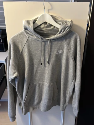 Grå hoodie från Nike med huva - Säljer en klassisk grå hoodie från Nike med broderad logga på bröstet. Hoodien har huva med dragsko och en stor magficka framtill. Tillverkad i mjukt bomullsmaterial som är perfekt för chill dagar eller när du vill ha en clean streetwear-look.