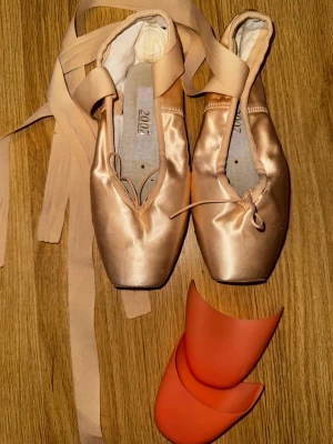 Rosa ballerinaskor i satin med tåskydd - Snygga ballerinaskor för balett i ljusrosa satin med långa band att knyta runt ankeln. Skorna har spetsig tå och platt sula, och kommer med två tåskydd i silikon för extra komfort. Perfekta för dig som dansar balett och vill ha klassiska tåskor.Freed of London Classic Professional